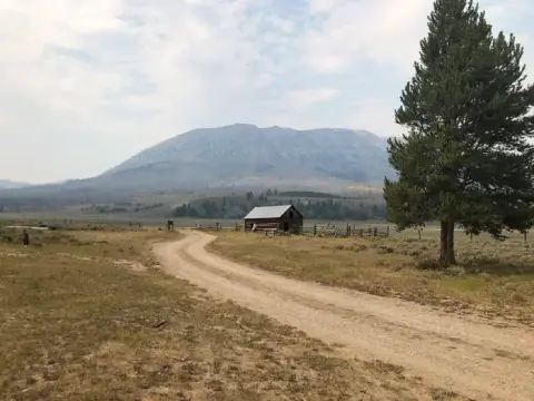A dirt road leads past a small wooden building toward a large mountain in a rural landscape.