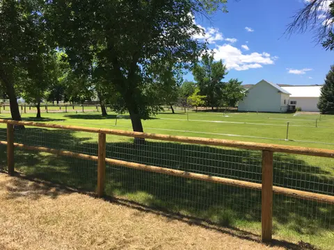 A wooden and wire fence borders a lush green lawn with sprinklers and white buildings under a blue sky.