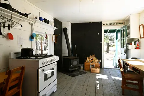 An interior view of a rustic cabin kitchen with a stove, wood-burning heater, and open doorway.