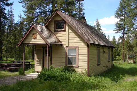 A small cabin stands among pine trees in a grassy clearing.