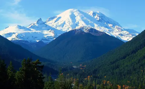 A snow-capped mountain rises behind dark forested hills under a blue sky.