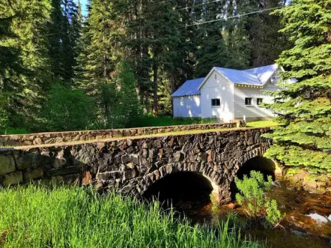 A stone bridge spans a small stream in front of a white building surrounded by a dense forest.