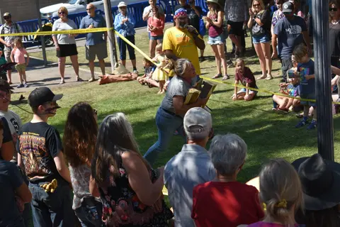 A woman carries logs at an outdoor community event with a crowd watching.