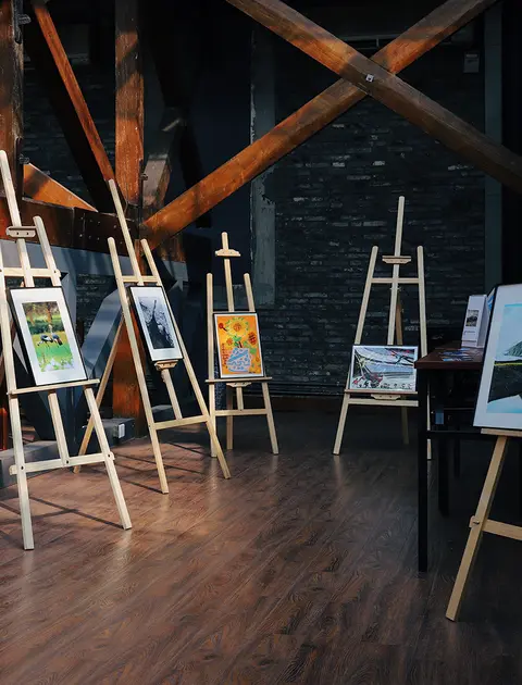 Several easels displaying artwork inside a room with wooden beams and a brick wall.