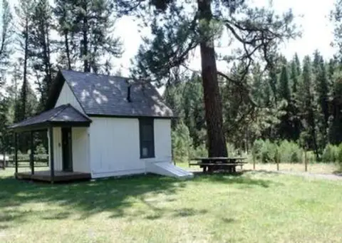 A small white cabin stands in a grassy clearing surrounded by pine trees.
