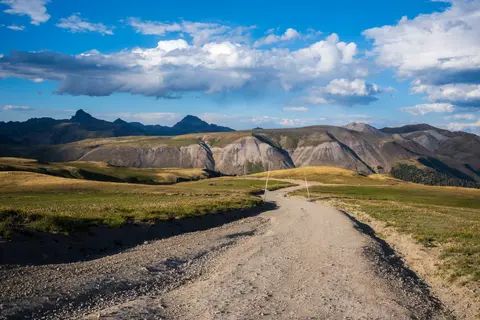 A dirt road winds through a mountainous landscape under a blue sky with fluffy clouds.