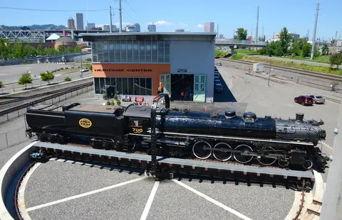 A large black steam locomotive sits on a turntable outside a heritage center building.