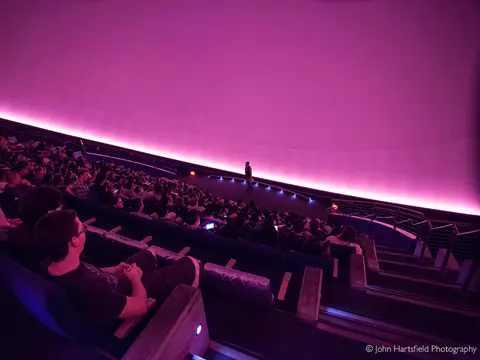 A large audience seated inside a domed auditorium with pink lighting.