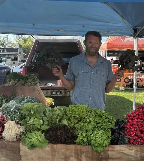 A smiling vendor holds up bunches of leafy greens at his farmers market stall.