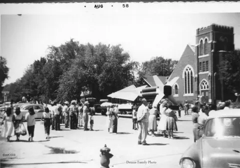 A historic black and white photo of people gathered on a street near a large building with a tower.