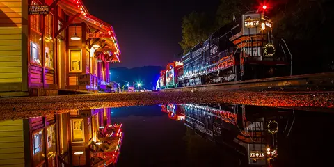 A train and a decorated station are illuminated at night with a reflection in a puddle.