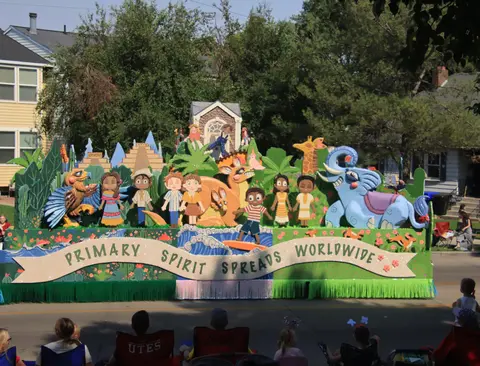 A colorful parade float featuring cartoon characters, animals, and the text 'PRIMARY SPIRIT SPREADS WORLDWIDE'.