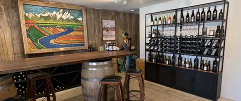 A rustic wine bar interior with a wooden counter, wine racks, and a large cork mosaic art piece.