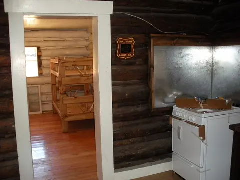 An interior view of a rustic log cabin showing a doorway to another room with bunk beds and a kitchen stove in the foreground.