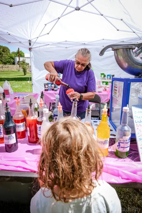 A woman pours syrup onto a snow cone at an outdoor stand while a child watches.