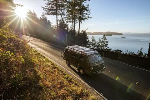 A camper van drives along a coastal road next to the ocean at sunrise.