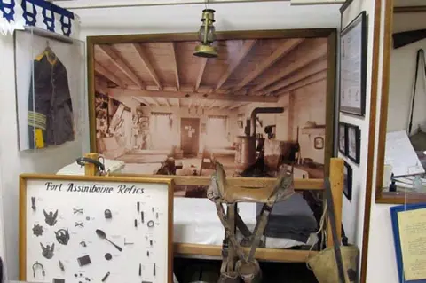 Interior exhibit featuring a large historic photo on the wall, a bed, a saddle, and a display case with artifacts.