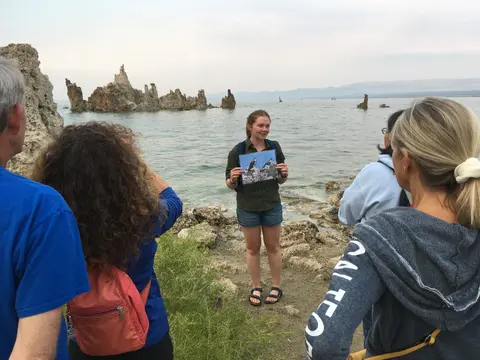 A guide holding a picture in front of a group of tourists near a lake with rock formations.