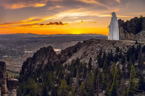 A large white statue stands on a mountain ridge overlooking a town at sunset.