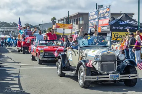 A street parade features classic cars and people waving American flags.