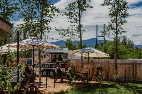 People sitting at outdoor picnic tables near a food truck with mountains and a bridge in the background.