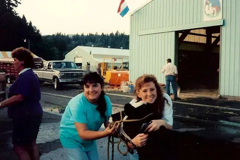 Two smiling young women pose with a calf near a barn and truck.