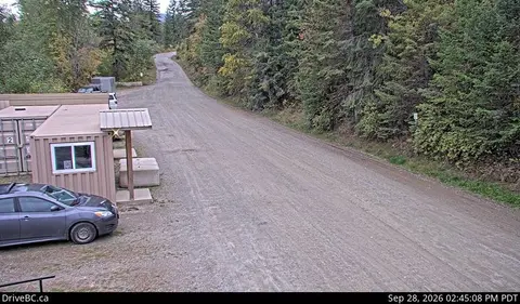 A dirt road next to a small building and a car, surrounded by forest.