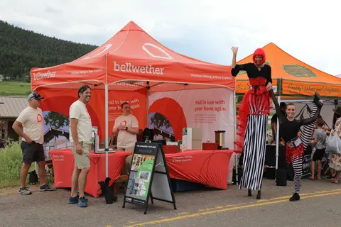 People stand near an outdoor promotional tent while a performer on stilts waves.