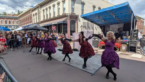 Performers in period costumes dance on a street stage during an outdoor event.