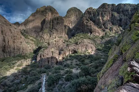 A rugged mountainous landscape with steep rocky slopes and sparse vegetation under a cloudy sky.