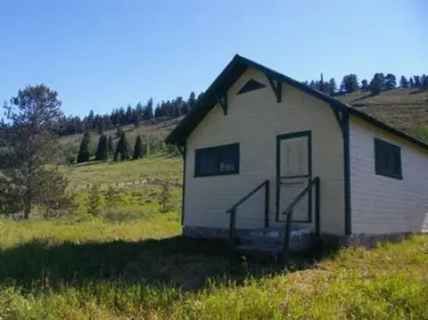 A small cabin stands in a grassy clearing with a hilly landscape in the background.