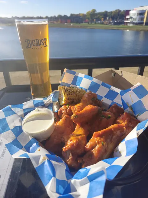 A basket of chicken wings, corn on the cob, dipping sauce, and a glass of beer are set on a table outdoors near water.