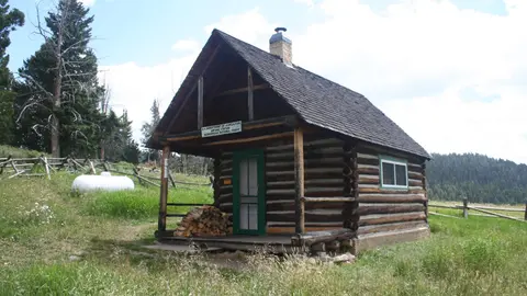 A log cabin sits in a grassy clearing with trees and a wooden fence nearby.