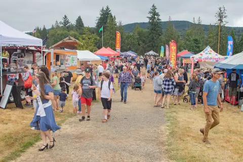 A crowd of people walking along a dirt path at an outdoor festival with food trucks and tents.