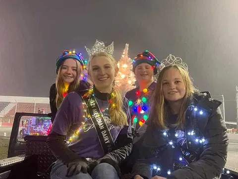 Four young people wearing crowns and holiday lights pose on a vehicle at night.