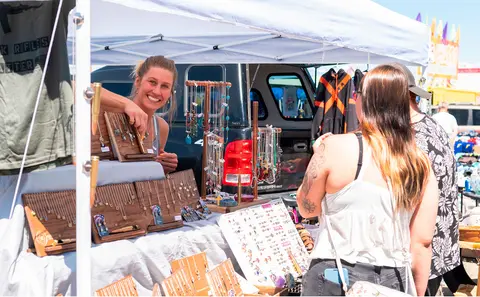 A smiling vendor shows jewelry at a market booth to a customer.