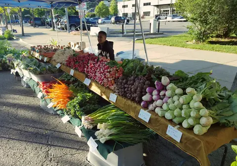 A vendor sits behind a table filled with fresh vegetables at an outdoor market.