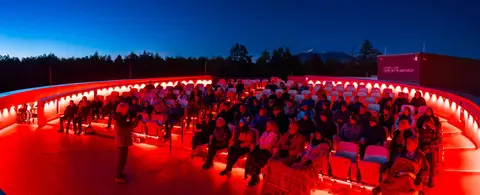 A speaker addresses a seated audience in an outdoor theater illuminated with red lighting at dusk.
