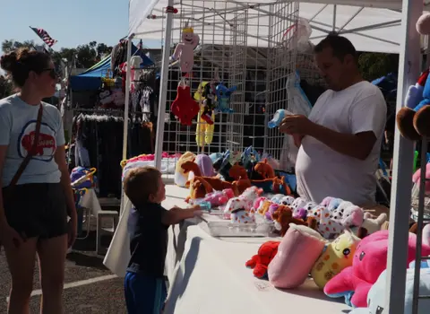 A vendor works at a booth filled with plush toys while a woman and child look at the merchandise.