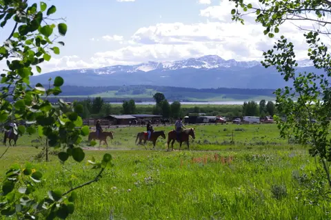 People ride horses across a green field with snowy mountains in the background.