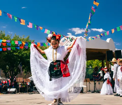 A young girl dances outdoors wearing a traditional white dress with a decorative skirt and colorful flags hanging overhead.