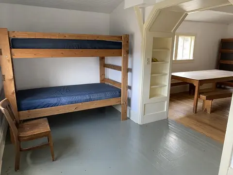 A rustic room features a wooden bunk bed and a chair, with a dining area visible through an arched doorway.