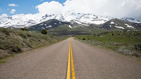 A paved road stretches toward snow-capped mountains under a partly cloudy sky.