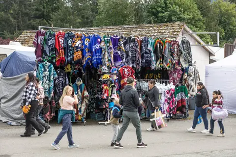 People walk past a market stall selling patterned blankets and apparel outdoors.