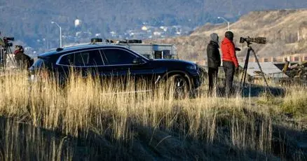 People stand near a vehicle and a camera on a tripod outdoors in a grassy area.