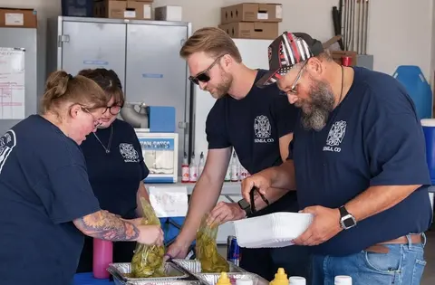 Four people in blue shirts are preparing food indoors.