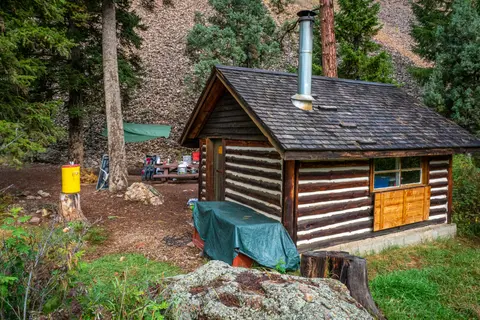 A small log cabin sits in a wooded area with a rocky slope in the background.