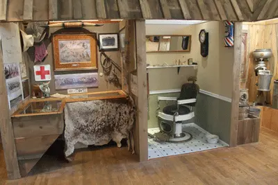 An indoor historical display showing a recreated vintage barbershop and a rustic room with various artifacts.