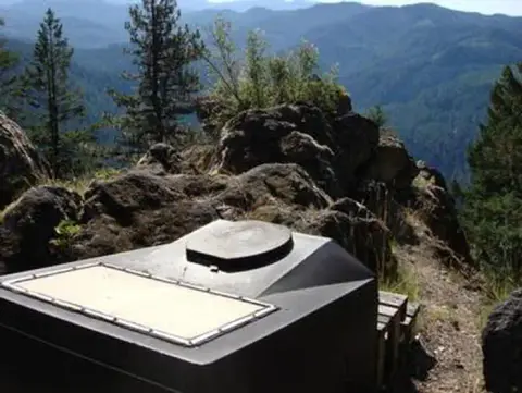A water tank or vault toilet sits on a rocky mountain overlook with forested hills in the background.