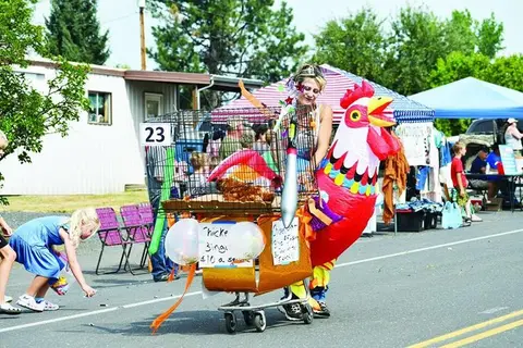 A woman pushes a cart with a large inflatable rooster and cage down a street during an outdoor event.